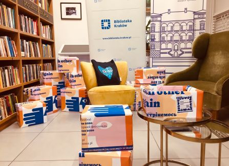 A photograph. the krakow library. all around the room cardboard boxes with books packed in colorful paper promoting the action multicultural library. In the background a bookshelf.